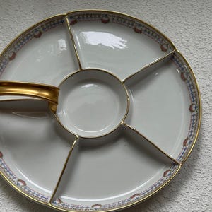 May include: A white porcelain serving platter with a gold handle and trim. The platter is divided into five sections, with a central circular compartment. The rim features a blue, red, and gold patterned border. A decorative piece for serving appetizers.