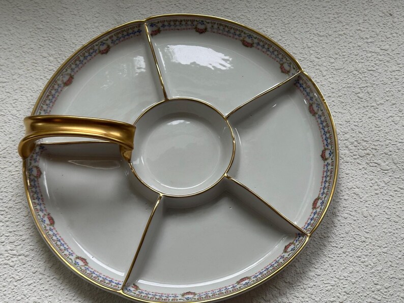 May include: A white porcelain serving platter with a gold handle and trim. The platter is divided into five sections, with a central circular compartment. The rim features a blue, red, and gold patterned border. A decorative piece for serving appetizers.
