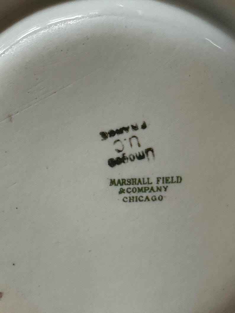 May include: Close-up of a white ceramic dish with black text. The text includes "U.C. France", "own", and "Marshall Field & Company Chicago". The dish has a slightly textured surface.