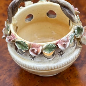 May include: A small, cream-colored ceramic basket with a twisted rope handle. The basket is decorated with pink and green flowers and has holes around the sides.
