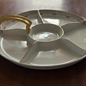 May include: A white porcelain serving platter with a gold handle and trim. The platter is divided into six sections, with a central round compartment and five surrounding sections. The rim features a decorative pattern in red, blue, and gold.