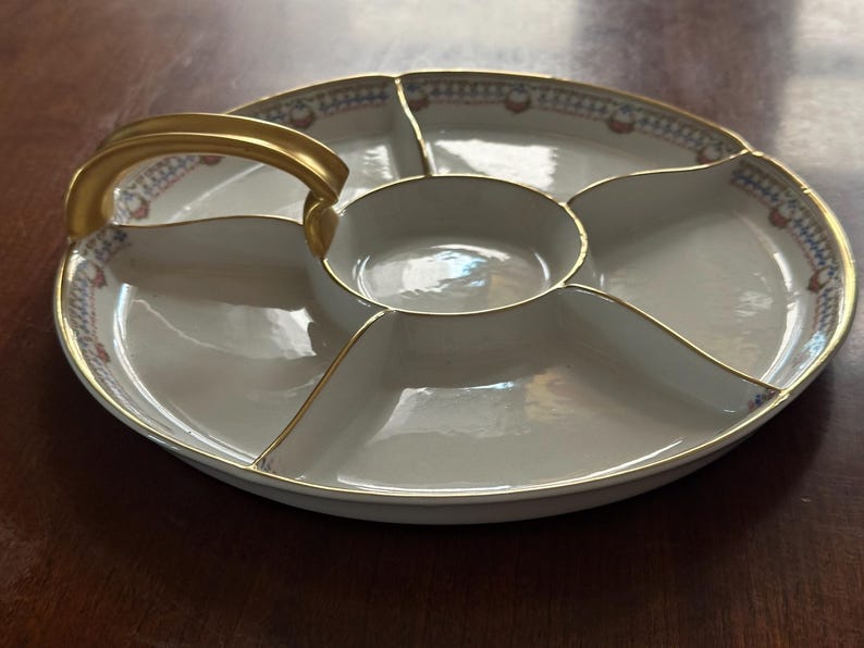 May include: A white porcelain serving platter with a gold handle and trim. The platter is divided into six sections, with a central round compartment and five surrounding sections. The rim features a decorative pattern in red, blue, and gold.