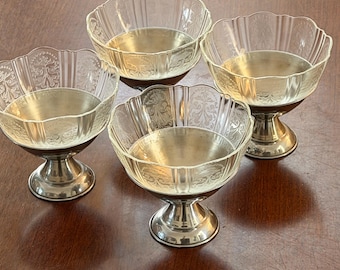 May include: A set of four clear glass dessert bowls with scalloped edges and etched floral patterns, each mounted on a polished silver-toned metal pedestal base. The bowls feature vertical ribbing and a wide, rounded opening for serving.