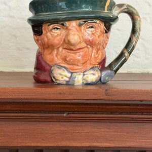 May include: A ceramic mug shaped like a smiling man's face, wearing a green hat and a bow tie with yellow accents. The mug has a brown handle with a marbled design and is set on a wooden surface.