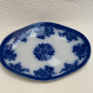 May include: An oval ceramic dish featuring a white base with a deep blue floral pattern. The design includes a prominent central floral cluster surrounded by smaller floral motifs distributed along the scalloped edges of the dish's rim.