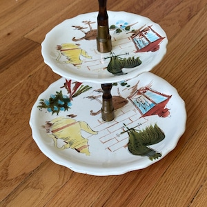 May include: Two-tiered ceramic serving stand with a wooden handle. The stand features a white background with hand-painted illustrations of chickens, roosters, and a brick wall.