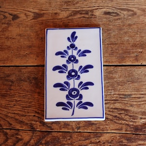 May include: A white ceramic tile with a blue floral design. The tile features a stylized plant with three blue flowers and leaves.
