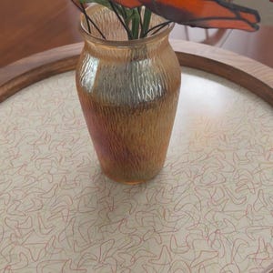 May include: A textured amber glass vase containing several stained glass poppy flowers with vibrant orange petals and green leaves. The vase has a tapered neck and a rippled surface, holding the wire-stemmed flowers in a decorative arrangement.