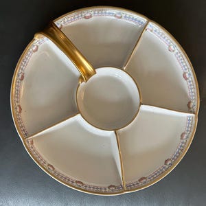 May include: A white porcelain serving platter with a gold handle and trim. The platter is divided into five sections, with a central circular well. The rim features a decorative pattern in blue, red, and gold.