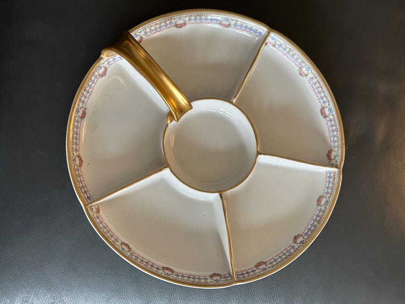 May include: A white porcelain serving platter with a gold handle and trim. The platter is divided into five sections, with a central circular well. The rim features a decorative pattern in blue, red, and gold.