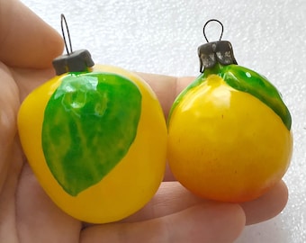 Op de afbeelding: Twee geel en groen citroenvormige kerstversieringen. Eén ornament heeft een groot groen bladontwerp, de andere een kleinere. Beide hebben een donkere metalen dop en een draadlus om op te hangen.