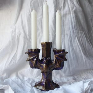 Puede incluir: Un candelero de tres brazos con un diseño de murciélago. El candelero está hecho de cerámica y está pintado en tonos de morado y dorado. Las velas son blancas.