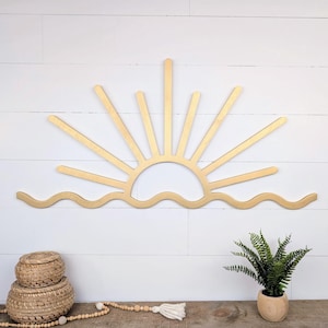 May include: A wooden sunburst wall art piece with a gold finish. The design features a sun with rays and a wavy line representing the ocean. The art is mounted on a white shiplap wall. Decorative baskets and a plant are on a wooden surface.