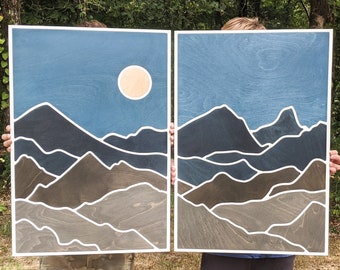 Puede incluir: Dos piezas rectangulares de arte mural con un diseño de paisaje montañoso. La obra de arte presenta un cielo azul con un sol beige y montañas en capas en tonos de gris y marrón, delineadas en blanco. Las piezas se sostienen frente a un fondo verde.