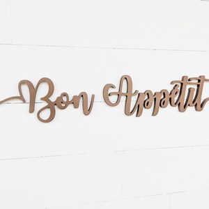 May include: A wooden sign with the words "Bon Appetit" in a cursive font. The sign is painted a light brown color and is mounted on a white wall.