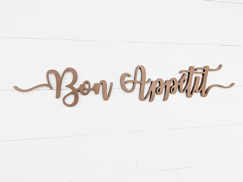 May include: A wooden sign with the words "Bon Appetit" in a cursive font. The sign is painted a light brown color and is mounted on a white wall.