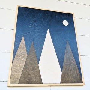 May include: A wooden wall art piece with a blue background and white mountains. The mountains are made of different shades of wood. There is a white circle in the upper right corner of the image, representing the moon.