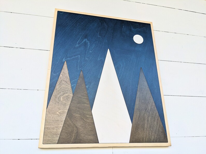 May include: A wooden wall art piece with a blue background and white mountains. The mountains are made of different shades of wood. There is a white circle in the upper right corner of the image, representing the moon.
