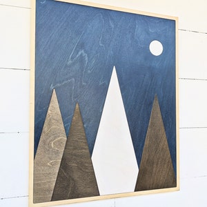 May include: A wooden wall art piece with a blue background and white mountains. The mountains are made of different shades of wood. There is a white circle in the upper right corner of the image.