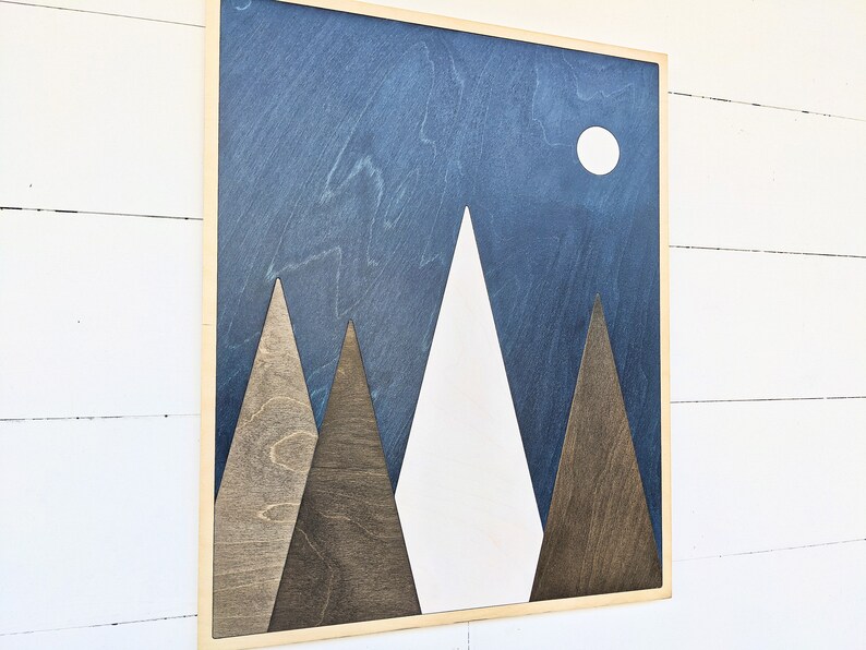 May include: A wooden wall art piece with a blue background and white mountains. The mountains are made of different shades of wood. There is a white circle in the upper right corner of the image.