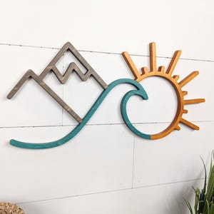May include: Wooden wall art featuring a mountain, wave, and sun design. The mountain is gray, the wave is teal, and the sun is orange. The artwork is mounted on a white, horizontal-planked wall. The overall theme is nature and coastal.