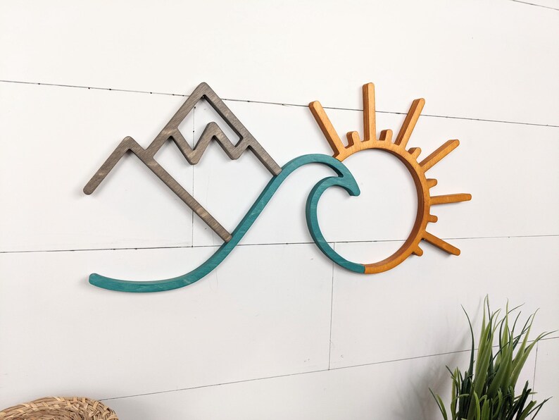May include: Wooden wall art featuring a mountain, wave, and sun design. The mountain is gray, the wave is teal, and the sun is orange. The artwork is mounted on a white, horizontal-planked wall. The overall theme is nature and coastal.