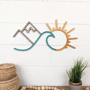 May include: Wooden wall art featuring a mountain, wave, and sun design. The mountain is gray, the wave is teal, and the sun is orange. This decorative piece is ideal for adding a coastal or nature-inspired touch to any room.