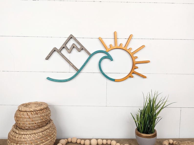 May include: Wooden wall art featuring a mountain, wave, and sun design. The mountain is gray, the wave is teal, and the sun is orange. This decorative piece is ideal for adding a coastal or nature-inspired touch to any room.