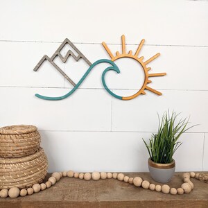 May include: Wooden wall art featuring a mountain range, a wave, and a sun. The mountain is gray, the wave is teal, and the sun is orange. The art is mounted on a white shiplap wall. Decorative items are in the foreground.