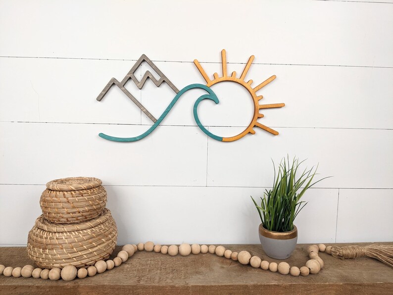 May include: Wooden wall art featuring a mountain range, a wave, and a sun. The mountain is gray, the wave is teal, and the sun is orange. The art is mounted on a white shiplap wall. Decorative items are in the foreground.