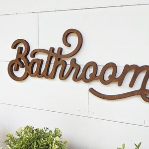 May include: Wooden "Bathroom" sign in a cursive font, stained a dark brown color. The sign is mounted on a white, horizontal shiplap wall. Greenery is visible in the bottom left corner.