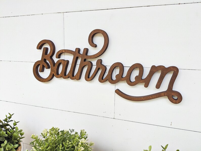 May include: Wooden "Bathroom" sign in a cursive font, stained a dark brown color. The sign is mounted on a white, horizontal shiplap wall. Greenery is visible in the bottom left corner.