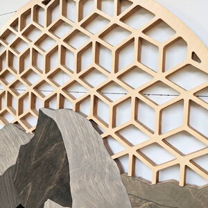 May include: A circular wooden wall art piece with a geometric pattern of overlapping hexagons. The bottom half of the piece features a layered mountain scene with a dark brown wood finish.