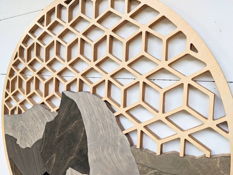 May include: A circular wooden wall art piece with a geometric pattern of overlapping hexagons. The bottom half of the piece features a layered mountain scene with a dark brown wood finish.