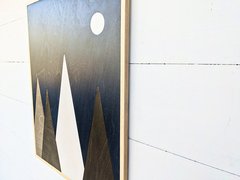 May include: A wooden wall art piece with a dark blue background and three white and brown triangles forming a mountain range. A white circle representing the moon is in the upper right corner.
