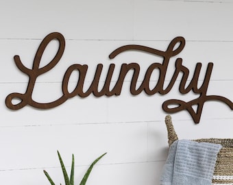May include: A wooden sign with the word "Laundry" in cursive script, mounted on a white wall. A small potted plant and a woven basket with a towel are on a wooden shelf below the sign.