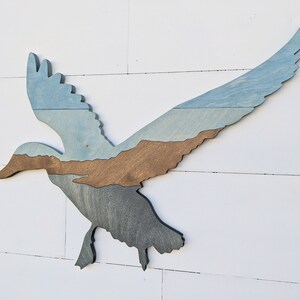 May include: A wooden silhouette of a flying duck with a blue, brown, and gray layered paint design.