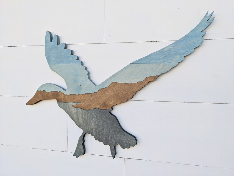 May include: A wooden silhouette of a flying duck with a blue, brown, and gray layered paint design.