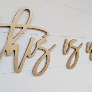 May include: Gold-colored wooden wall art spelling out "This is Us" in a cursive font. The letters are mounted on a white, horizontal-planked wall. The art is a decorative piece for home decor.