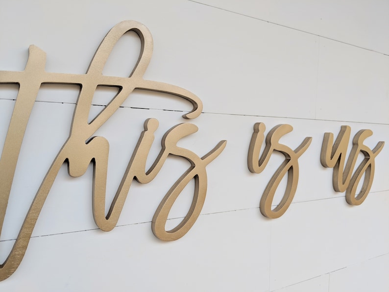 May include: Gold-colored wooden wall art spelling out "This is Us" in a cursive font. The letters are mounted on a white, horizontal-planked wall. The art is a decorative piece for home decor.