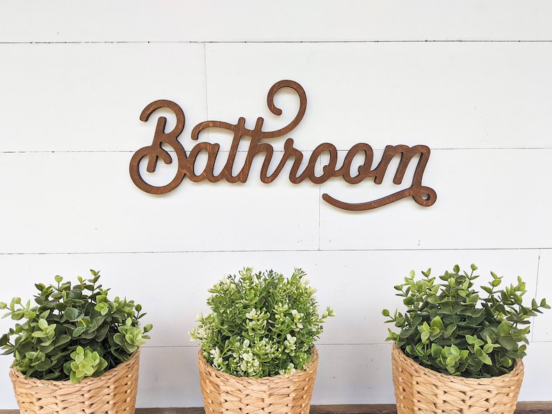 May include: Wooden "Bathroom" sign in a cursive font, mounted on a white shiplap wall. Below the sign are three small potted plants in woven baskets, adding a touch of greenery to the decor.