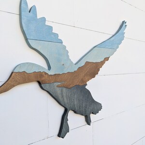 May include: A wooden wall decor in the shape of a flying duck. The duck is painted in shades of blue, brown, and gray, creating a layered effect.