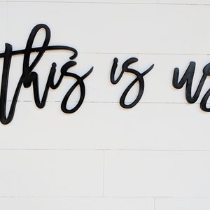 May include: Black wooden wall art spelling out "this is us" in a cursive font. The letters are connected and mounted on a white, horizontal plank background. A decorative piece for home decor.