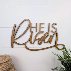 May include: Wooden sign with the words "He is Risen" in a cursive font. The sign is made of a light brown wood and is displayed against a white background. A woven basket and a green plant are in the foreground.