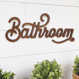 May include: Wooden "Bathroom" sign in a cursive font, stained a warm brown. The sign is mounted on a white, horizontal shiplap wall. Green plants in small pots are visible at the bottom of the image.