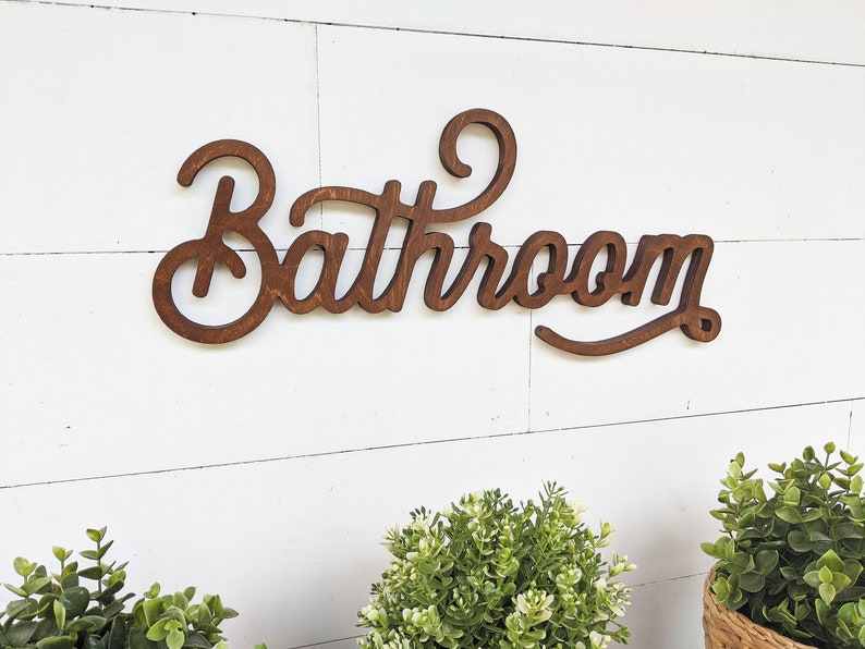 May include: Wooden "Bathroom" sign in a cursive font, stained a warm brown. The sign is mounted on a white, horizontal shiplap wall. Green plants in small pots are visible at the bottom of the image.