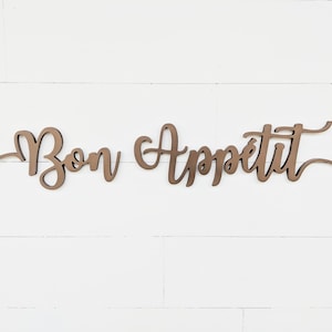 May include: A wooden sign with the words "Bon Appetit" written in a cursive font. The sign is painted in a brown color and is mounted on a white wall.