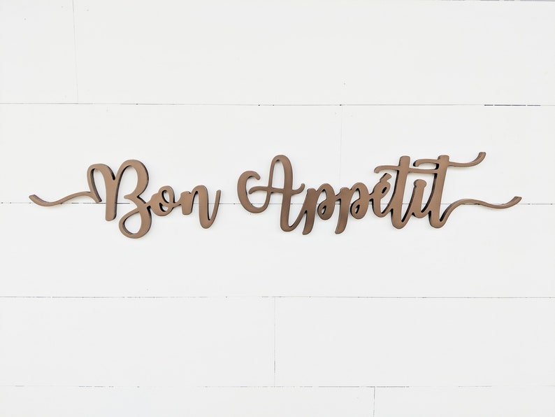 May include: A wooden sign with the words "Bon Appetit" written in a cursive font. The sign is painted in a brown color and is mounted on a white wall.