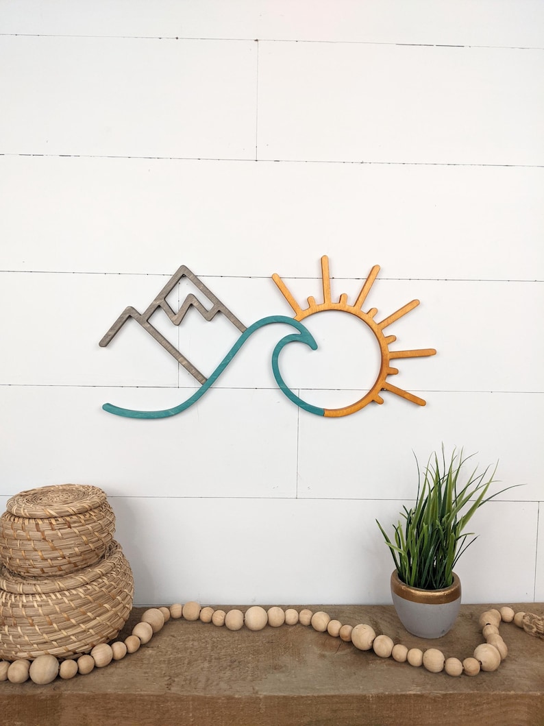 May include: Wooden wall art featuring a mountain range, a wave, and a sun. The mountain is gray, the wave is teal, and the sun is orange. The art is mounted on a white shiplap wall. Decorative items are in the foreground.