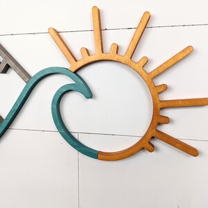 May include: Wooden wall art featuring a mountain range, a teal wave, and a sunburst design. The mountain is gray, the wave is teal, and the sun is a warm wood tone. The art is mounted on a white, horizontal-planked wall.